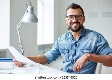 Smiling businessman in denim shirt and eyeglasses looking at camera in office