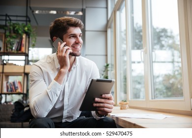 Smiling businessman dressed in white shirt sitting in cafe and using tablet computer while talking by his phone. Look aside.