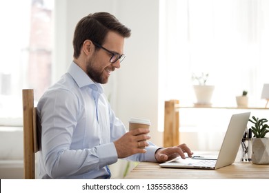 Smiling businessman drinking coffee and using laptop at workplace, confident with cardboard cup, typing on keyboard, looking at screen, reading business email, chatting with friends during break - Shutterstock ID 1355583863