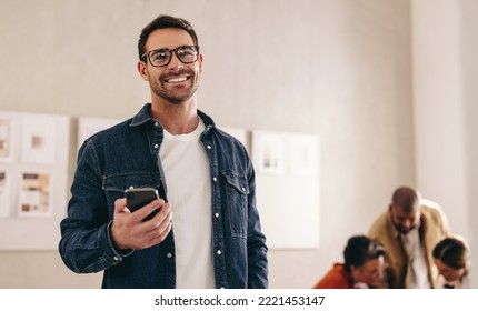 Smiling businessman with eyeglasses holding a smartphone while standing in an office. Happy young businessman working in a creative and modern workplace with his colleagues. - Powered by Shutterstock