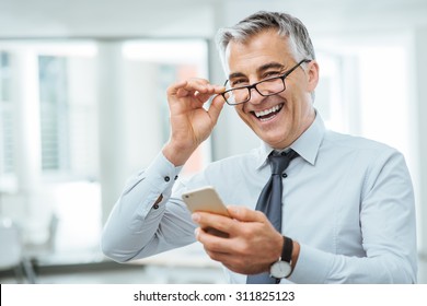 Smiling Businessman With Eyesight Problems, He Is Adjusting His Glasses And Reading Something On His Mobile Phone