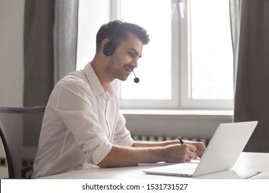 Smiling Businessman In Headset Using Laptop, Looking At Screen, Employee Student In Headphones With Microphone Watching Webinar, Making Notes, Studying Online, Interpreter Translating Course