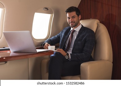smiling businessman holding cup of coffee in plane with laptop during business trip  - Shutterstock ID 1532582486