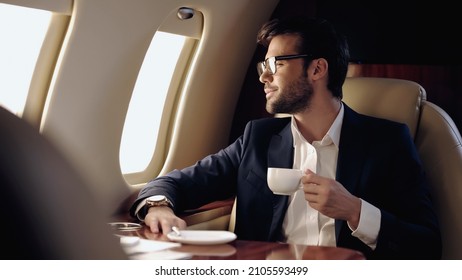 Smiling businessman holding cup and looking at window in private plane - Shutterstock ID 2105593499