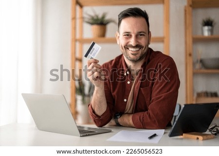 Smiling businessman holding and showing credit card while sitting in home office. Male business manager advertising online internet banking services, loan or deposits, cashback for purchases concept.	