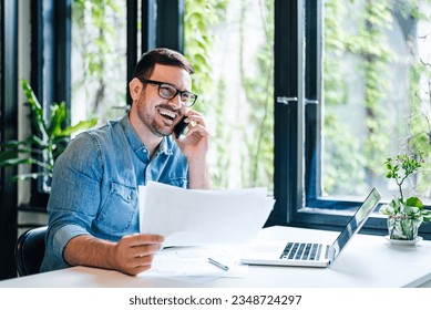 Smiling businessman with laptop discussing on smart phone. Male executive multitasking while working with documents at table. He is wearing casuals while sitting at home office. - Powered by Shutterstock