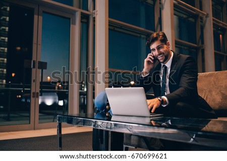 Smiling businessman with laptop talking on cellphone at the airport waiting lounge. Handsome man at waiting room in airport terminal.