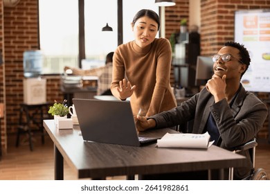 Smiling businessman listening as his asian coworker explains market trends using digital chart on laptop. Diverse team brainstorming strategy ideas, reviewing financial analytics on an office desk.
