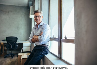 Smiling businessman looking at camera as he stands by a window sill with arms crossed. Mid adult businessman at his office. - Shutterstock ID 1414526951
