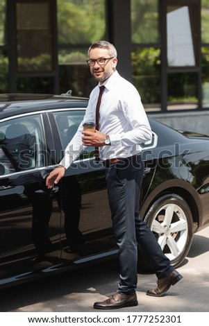 Smiling businessman looking at camera while holding paper cup near auto on urban street