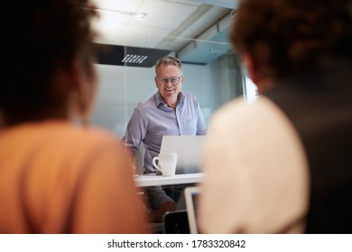 Smiling Businessman Looking At Colleagues Through Glass Shield Working In Creative Office During COVID-19