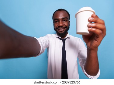 Smiling Businessman Raising Cup Of Coffee To Go At Camera In Videocall Conference On Blue Background. Wide Angle Pov Selfie Of Happy Vlogger Showing Styrofoam Container With Lid For Social Media.