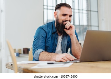 Smiling businessman resting his chin on his hand reading an online report on his laptop computer at a table in the office - Shutterstock ID 1038742600