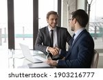 Smiling businessman shaking client hand, closing successful deal, sitting at table with laptop in office, satisfied hr manager hiring new employee, business partners handshaking at meeting