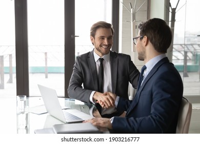 Smiling businessman shaking client hand, closing successful deal, sitting at table with laptop in office, satisfied hr manager hiring new employee, business partners handshaking at meeting