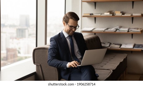 Smiling businessman sit on couch at office coworking area hold video conversation using laptop. Happy young man ceo executive work on pc online at corporate network glad to find decision solve problem - Shutterstock ID 1906258714