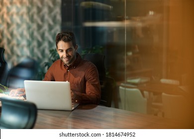 Smiling businessman sitting alone at his desk and using a laptop while working late in a dark office