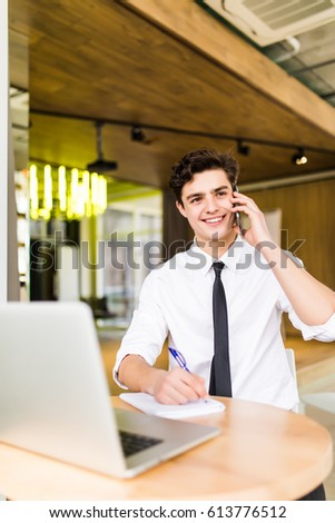 Smiling businessman sitting behind his desk with laptop and talking on mobile phone in office