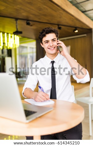 Smiling businessman sitting behind his desk with laptop and talking on mobile phone in office