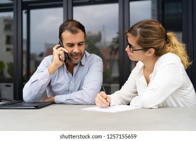 Smiling businessman sitting at meeting at talking on phone, young Caucasian businesswoman wearing glasses writing after him. Cooperation concept