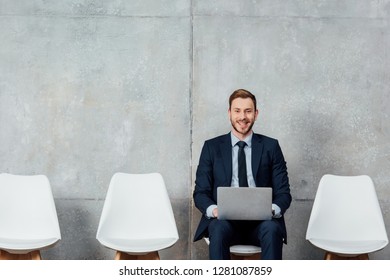 smiling businessman sitting on chair and using laptop in waiting hall