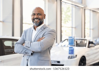 Smiling Businessman Standing With Arms Crossed At New Car Showroom. Confident Senior Car Dealer In Dealership While Looking At Camera. Portrait Of Professional Black Salesman In Luxury Auto Showroom.