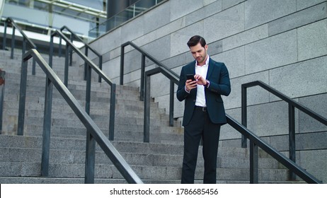 Smiling Businessman Standing With Smartphone Outdoors. Attractive Business Man Using Phone At Modern Street. Confident Businessman Checking Time On Wristwatch Outside.