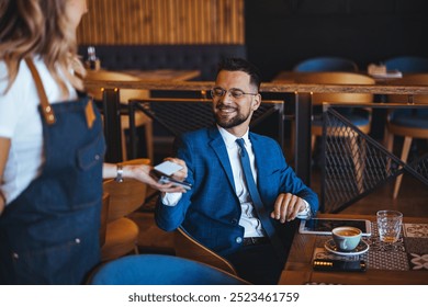 A smiling businessman in a suit pays for coffee using a credit card in a contemporary cafe setting, showcasing modern dining and payment methods. - Powered by Shutterstock