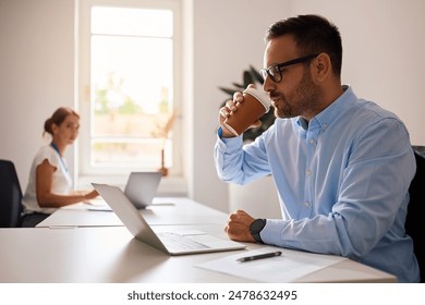 Smiling businessman using laptop and drinking coffee while working in the office. Copy space.  - Powered by Shutterstock