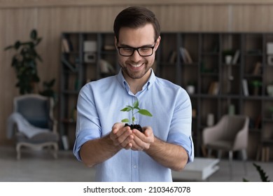 Smiling businessman wearing glasses holding small green plant sprout with soil, standing, happy entrepreneur employee with growing tree, startup project, profit, investment and growth concept - Powered by Shutterstock