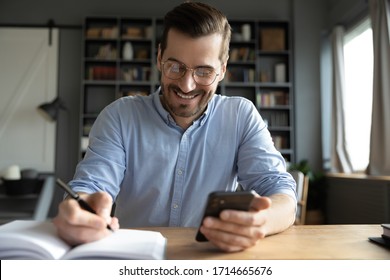 Smiling businessman wearing glasses using phone, writing important information in notebook, sitting at desk, happy man holding smartphone, making notes in personal daily planner, planning work day - Shutterstock ID 1714665676