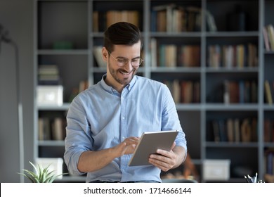 Smiling businessman wearing glasses using computer tablet, standing in modern cabinet, happy positive man enjoying leisure time, playing game, chatting in social network, browsing apps