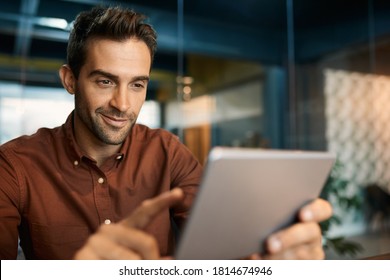 Lächelnder Geschäftsmann, der am frühen Abend allein an seinem Schreibtisch in einem Büro arbeitet und eine digitale Tablette verwendet