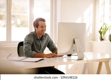 Smiling Businessman Working On Computer. Man Sitting At His Work Table Working On Computer At Home Listening To Audio Using Earphones.