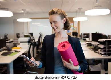 Smiling businesswoman with exercise mat using mobile phone at office - Powered by Shutterstock