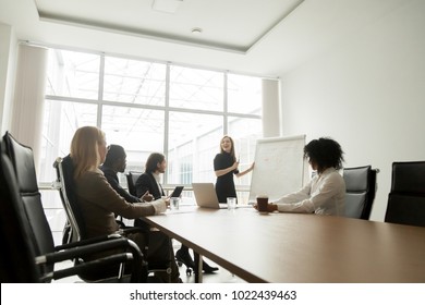 Smiling businesswoman giving presentation to diverse partners in meeting room, positive business coach explaining new project to multiracial group, team leader presenting marketing plan on whiteboard