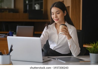 Smiling businesswoman holding coffee cup and working on laptop computer.