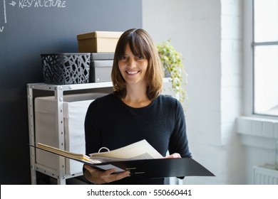 Smiling businesswoman holding an open binder full of paperwork in her hands as she stands in the office with a happy satisfied look - Shutterstock ID 550660441