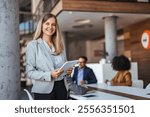 Smiling businesswoman holding a tablet in a modern office setting, conveying professionalism and teamwork with colleagues in the background. Ideal for corporate, workplace, and professional themes.