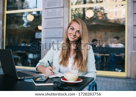 Smiling businesswoman with long hair writing on diary with pen while sitting at table with open netbook and cup of coffee in street cafeteria