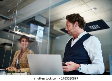 Smiling Businesswoman Looking At Businessman Through Glass Shield In Office During COVID-19