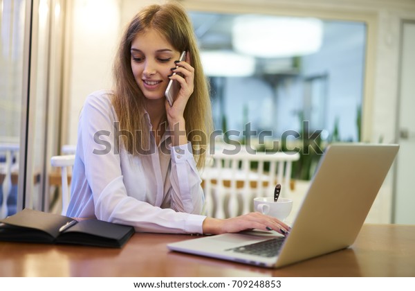 Smiling businesswoman making reservation online\
while talking on mobile telephone with assistant, female\
entrepreneur banking via laptop confirming payment communicating\
with service operator