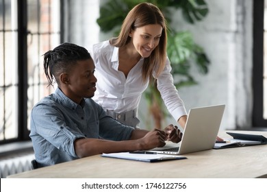 Smiling businesswoman mentor helping new African American employee with corporate software, training and teaching, standing near desk, looking at screen, supervisor checking intern work results
