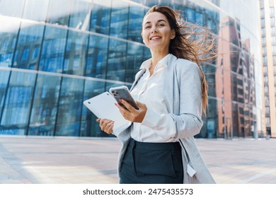 Smiling businesswoman on a phone call, holding a laptop, standing in front of a modern glass building.

 - Powered by Shutterstock