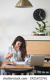 Smiling Businesswoman Reading Business Papers And Drinking Coffee. Business Woman Working From Home, Sitting At Desk With Laptop Computer, Digital Papers And Business Documents