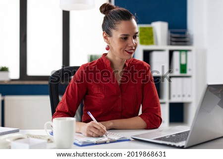 Smiling businesswoman reading data from laptop screen and taking notes on clipboard. Manager doing administrative wwork. Business leader doing financial expertise for client.