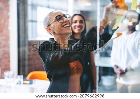 Smiling businesswoman sharing her ideas with her colleagues in a creative workplace. Confident young businesswoman sticking adhesive notes to a glass wall with her team standing in the background.