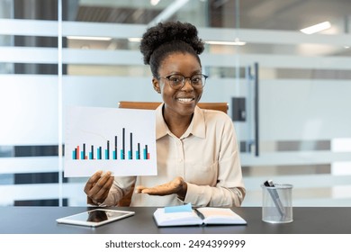 Smiling businesswoman showing financial chart during office meeting. Confident professional explaining business performance with graph. Modern office environment with tablet and documents on desk. - Powered by Shutterstock