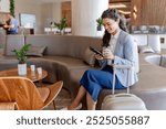 A smiling businesswoman sits in a modern hotel lobby, checking her phone while holding onto her suitcase. She appears relaxed and ready for her trip, surrounded by a comfortable lounge area.