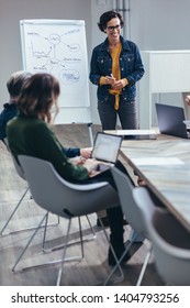 Smiling Businesswoman Standing In Front Of Flip Chart For Presentation With Team. Woman Giving Presentation On Sales Strategies To Colleagues In Board Room.